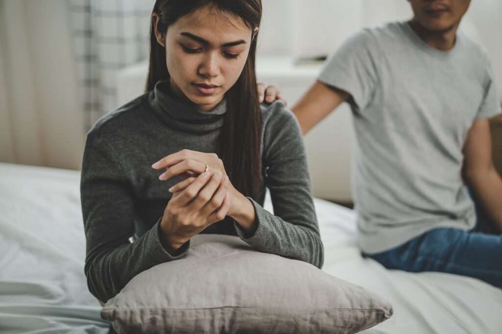 A woman looking at her wedding ring on the bed. Her husband is putting his hand on her shoulder. They are really struggling with being in an unequally yoked marriage.