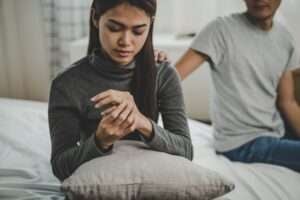 A woman looking at her wedding ring on the bed. Her husband is putting his hand on her shoulder. They are really struggling with being in an unequally yoked marriage.