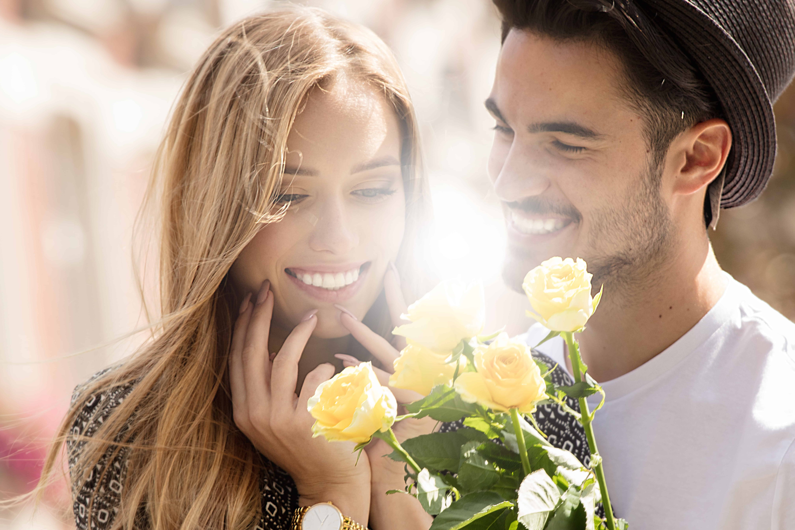 a guy giving a girl yellow roses looking at her while she smiles excited to receive the flowers.