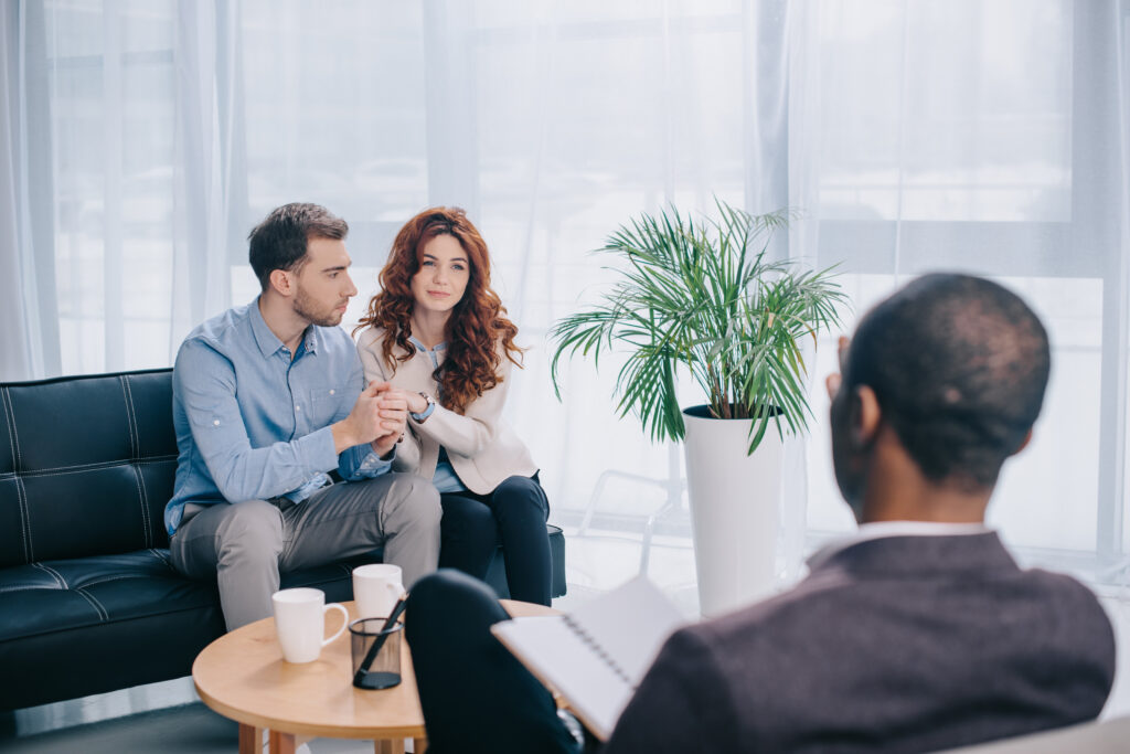A young couple sitting down trying to improve their communication skills with a black christian marriage counselor, holding a notebook and a pen.