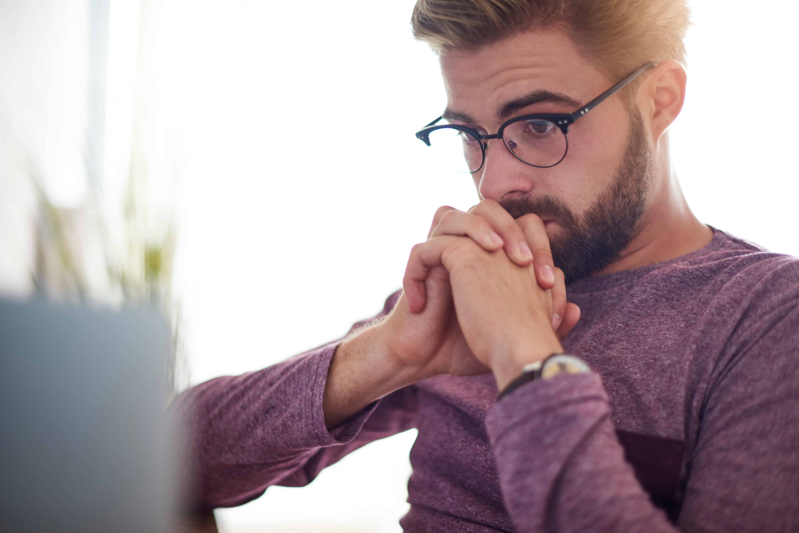 A guy wearing glasses with his hands clasped together looking at his computer screen.