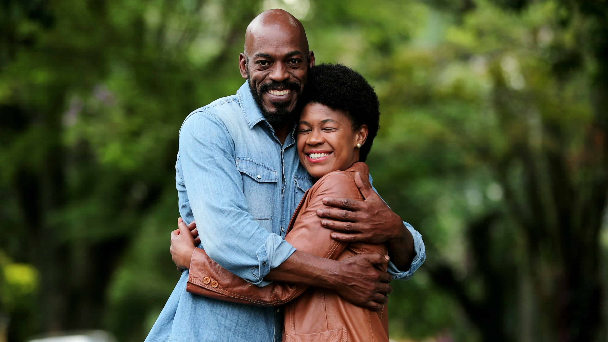 A black couple outside surrounded by trees hugging each other tightly.