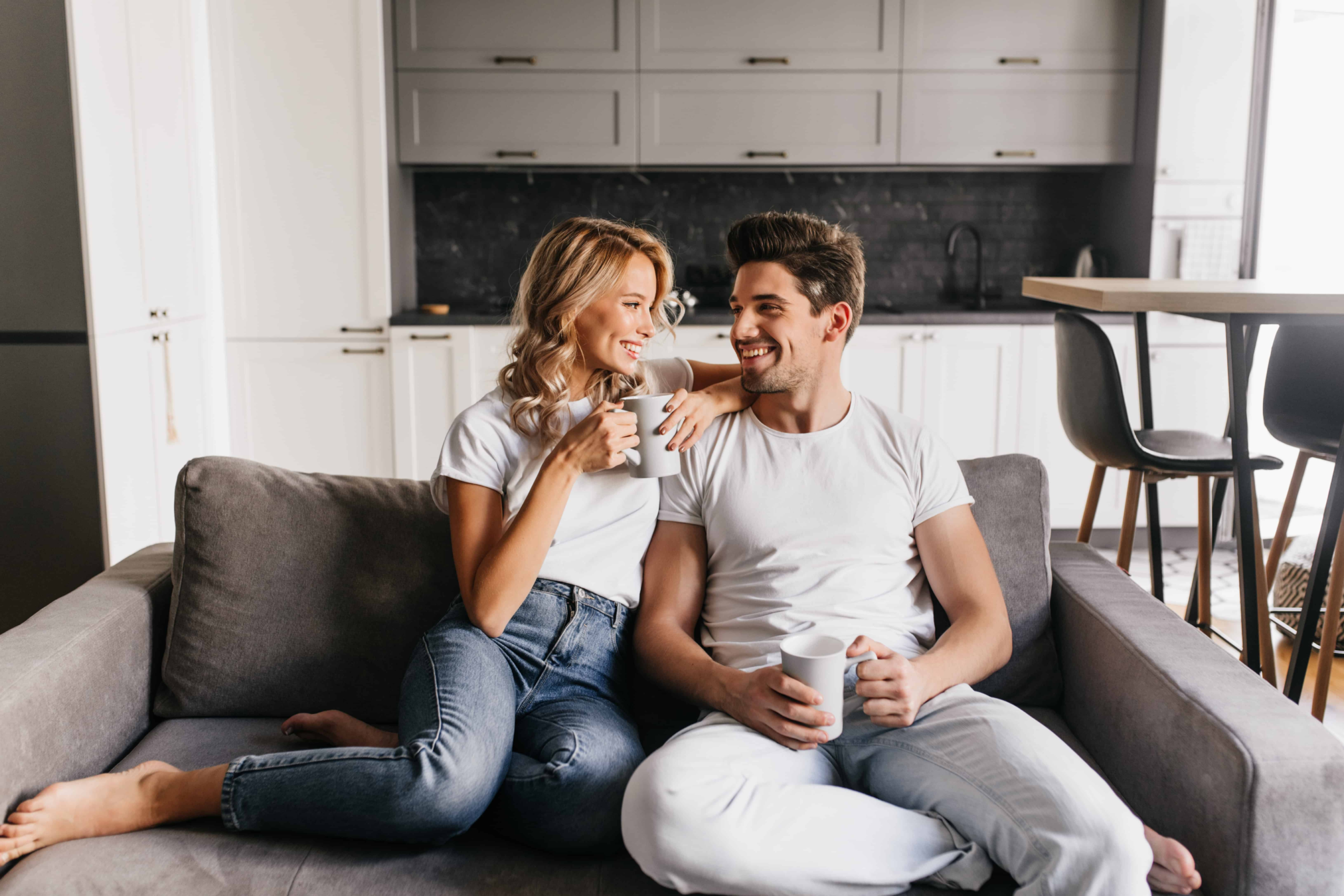 Couple sitting on a loveseat drinking coffee as they stare at one another.