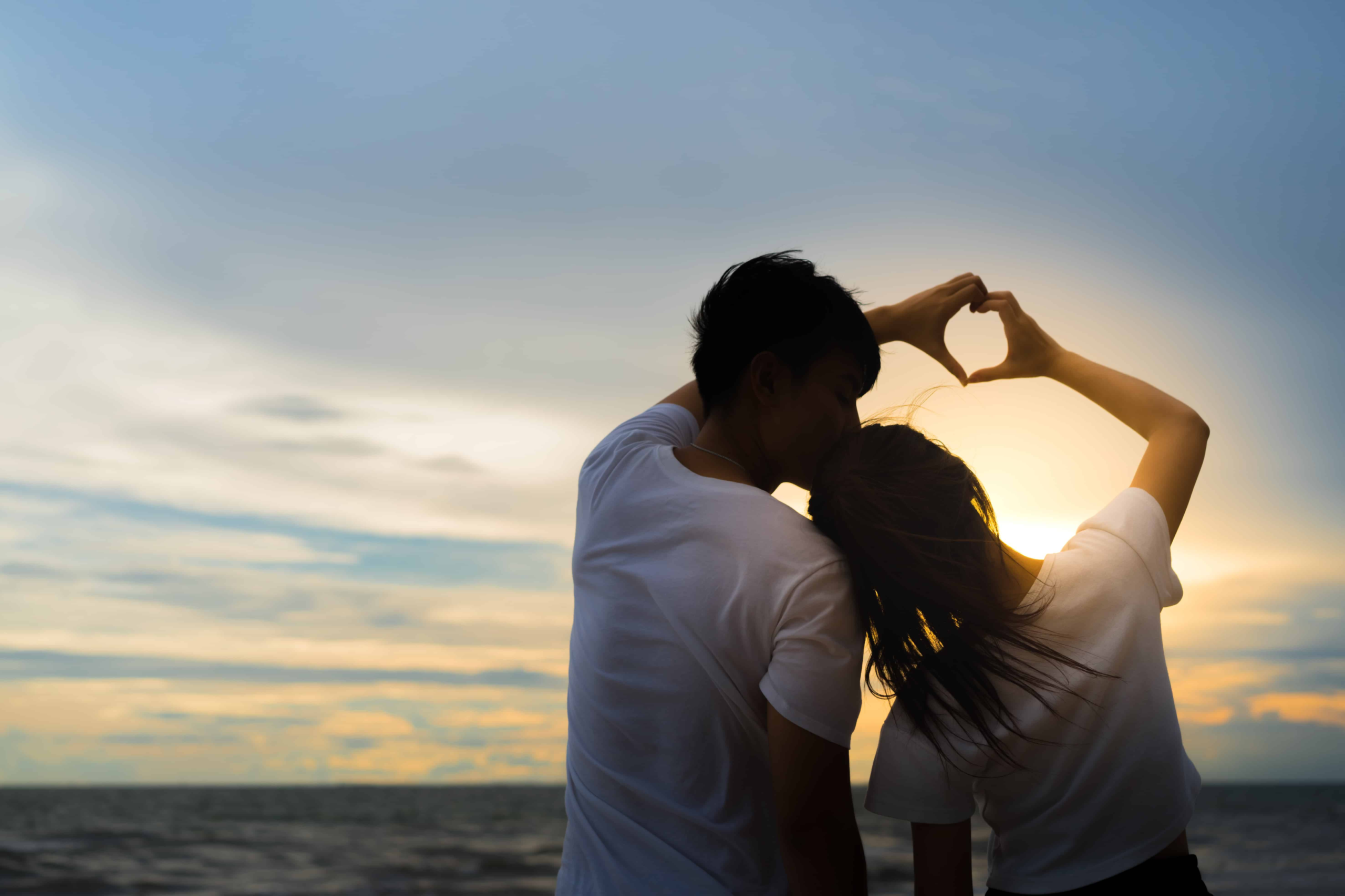 A beautiful beach is the background as the couple makes a heart with their hands. Only their silhouette can be seen.