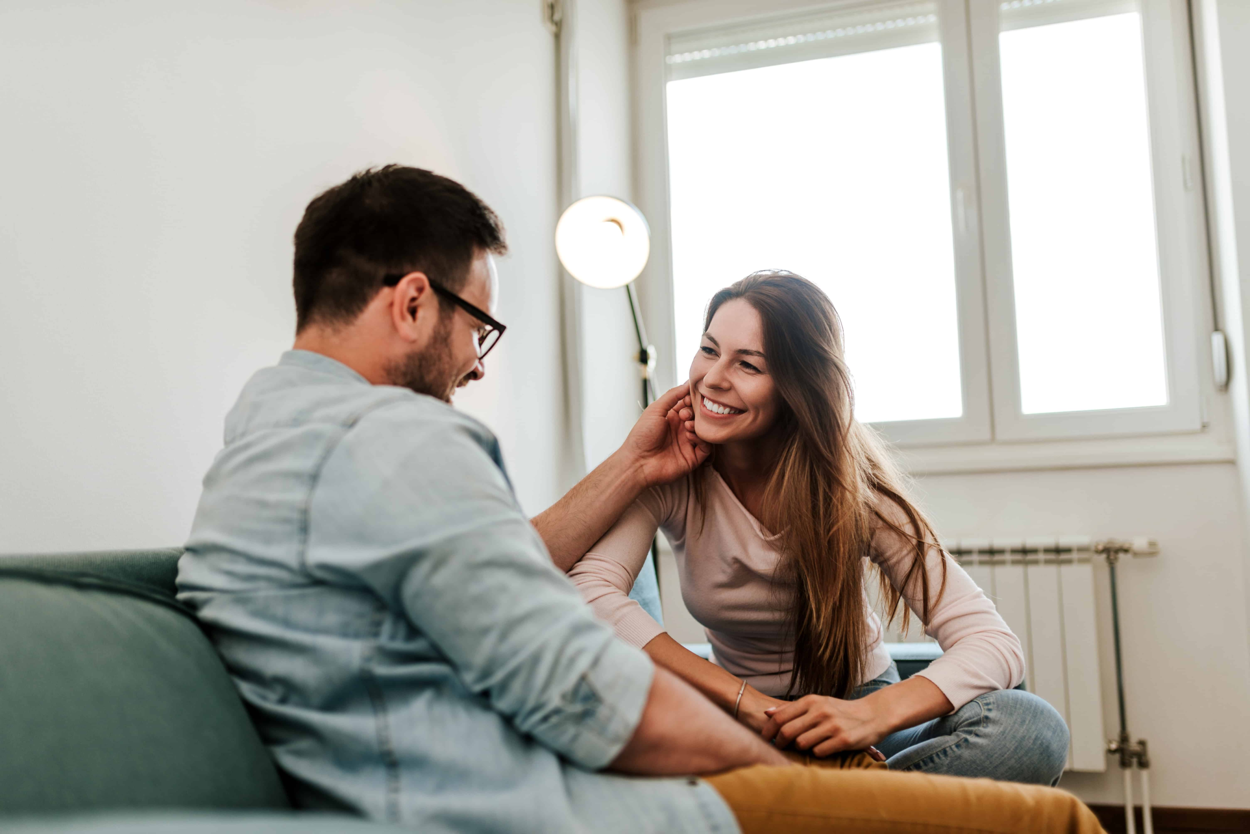 a man and a woman sitting on a green couch together. The man is rubbing his wife's face while the woman leans in intently. The woman is showing us how to prioritize my husband through her engaged body language.