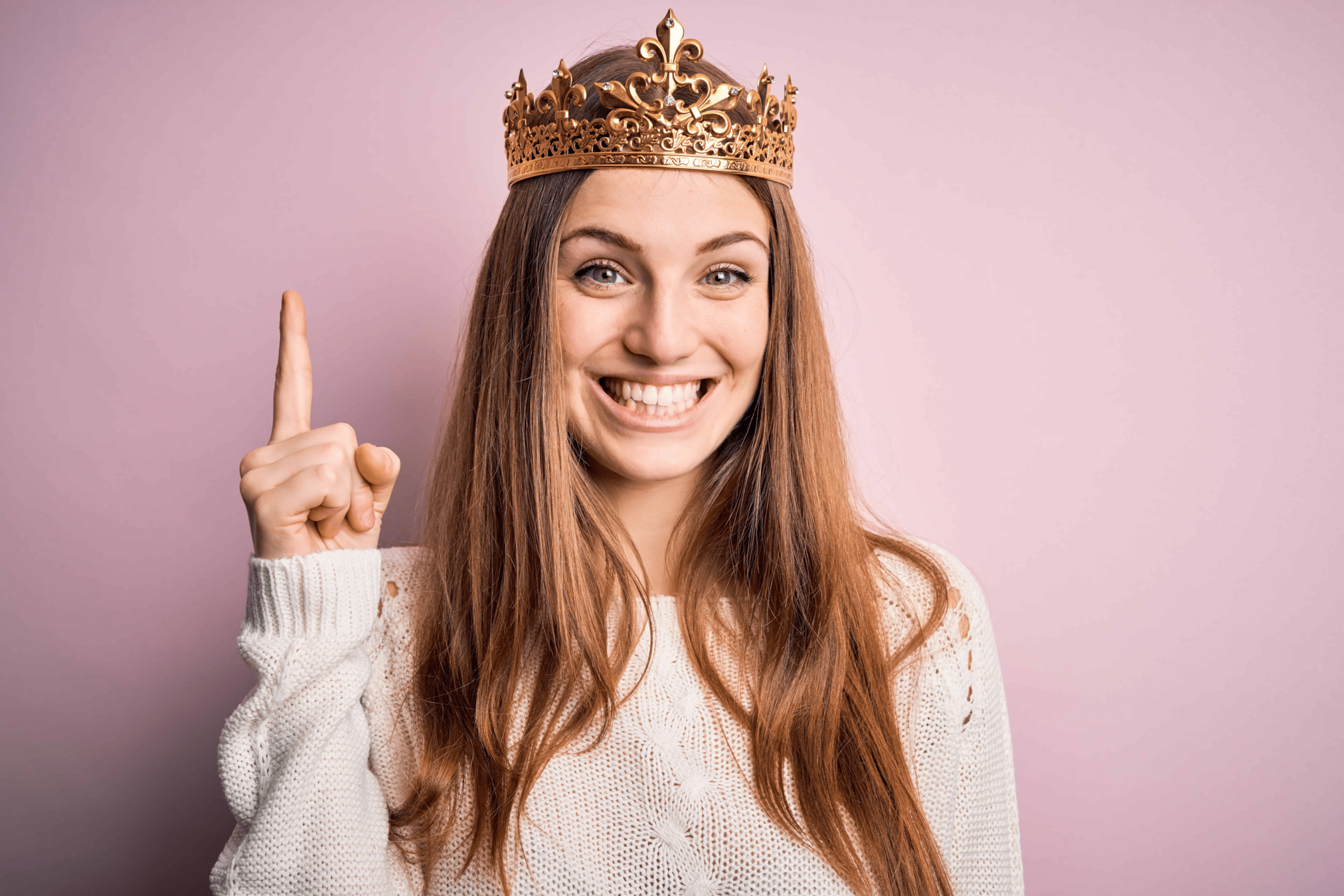 A woman with a crown on her head pointing upwards with a big smile on her face.