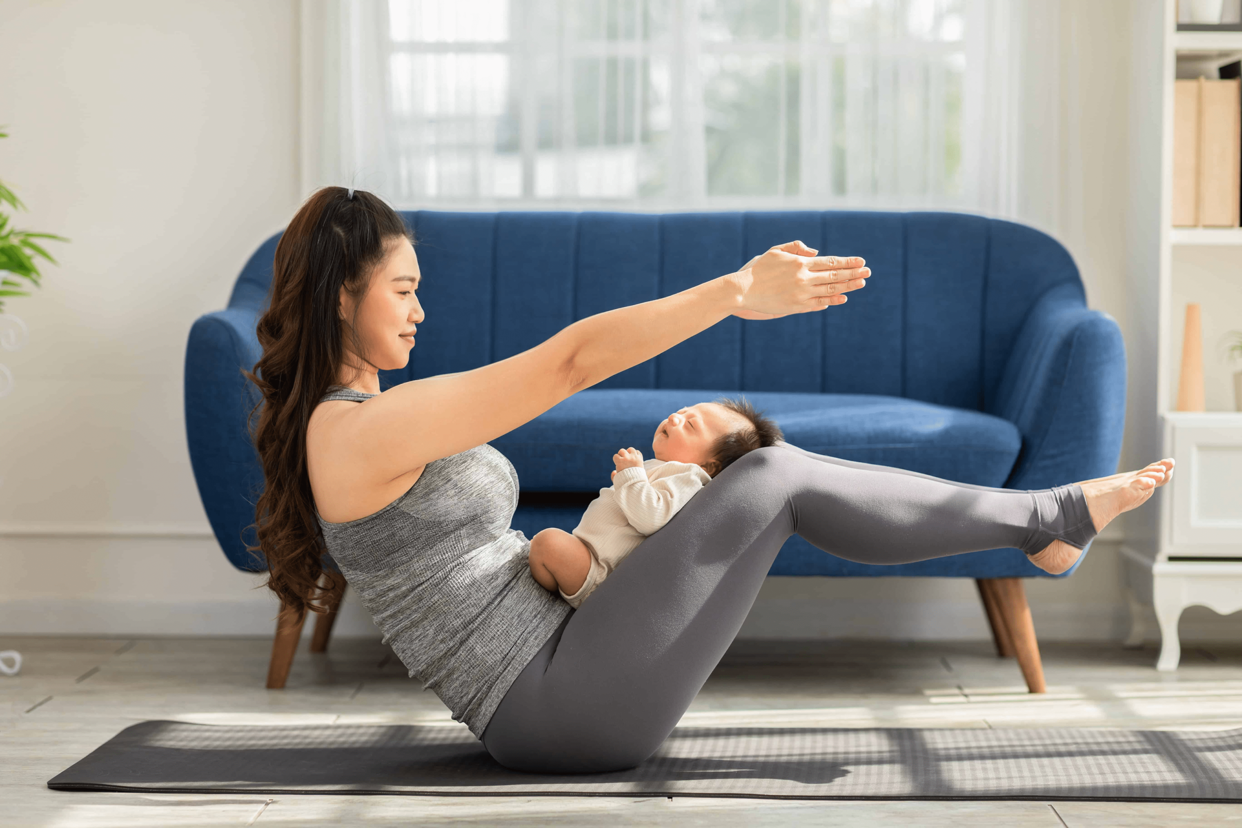 An asian woman sitting in her living room with a couch behind her and a yoga mat underneath her. The woman is doing a yoga pose while she has her baby on her lap.