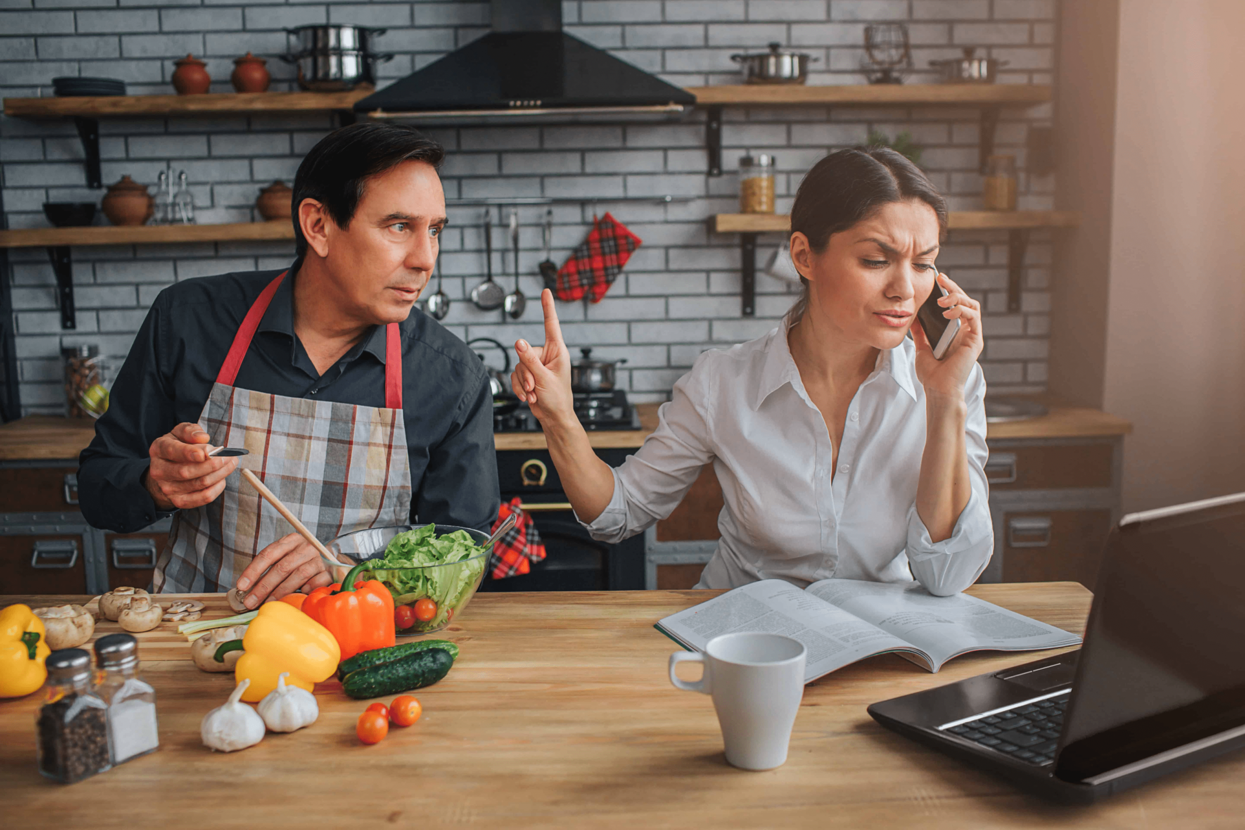 A man and woman in a kitchen cooking. The man wants the woman's attention but she is saying hold on as she is on the phone and on the computer at the same time.