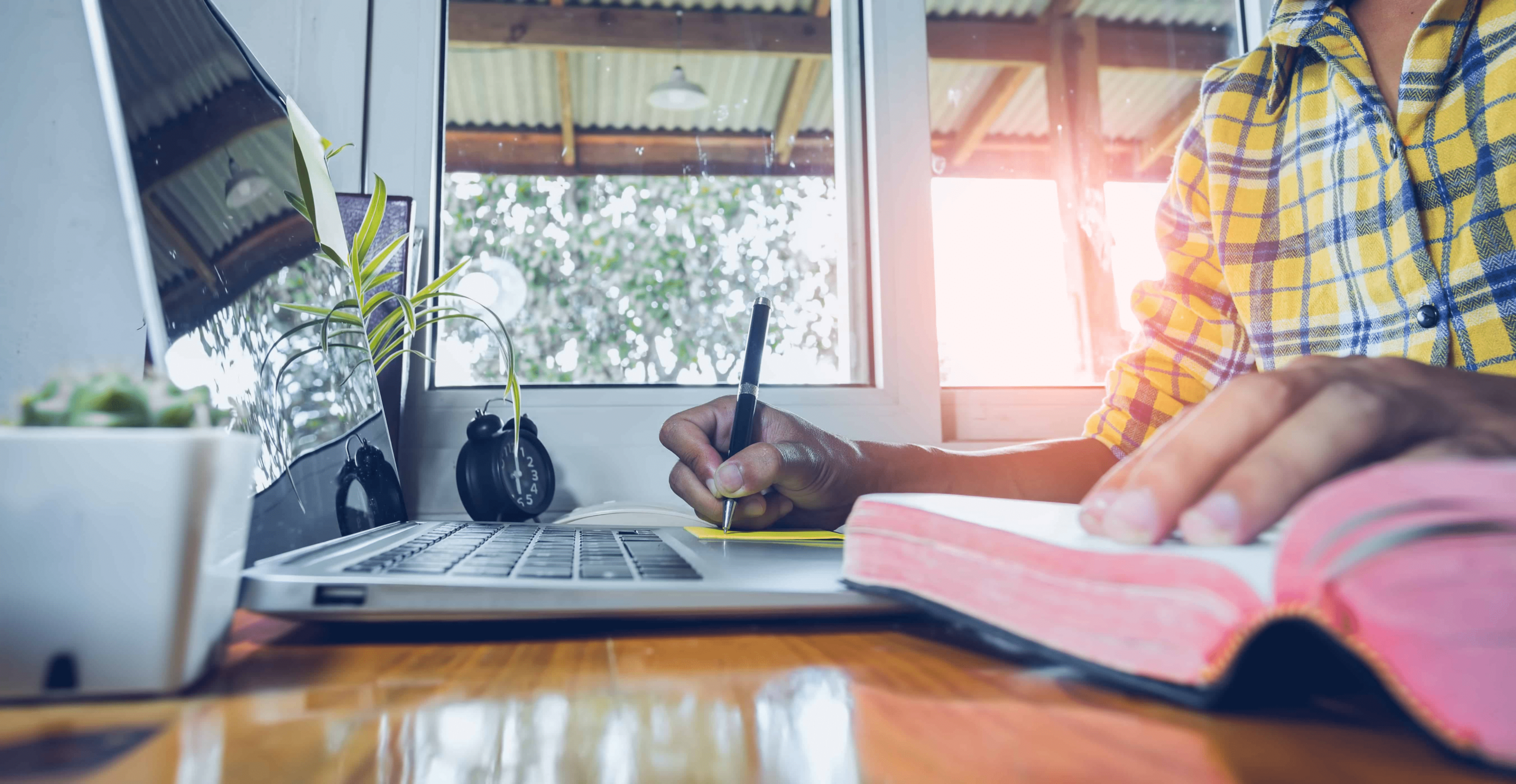 A man reading a book with his computer, book and journal in front of him. He is writing in his journal.