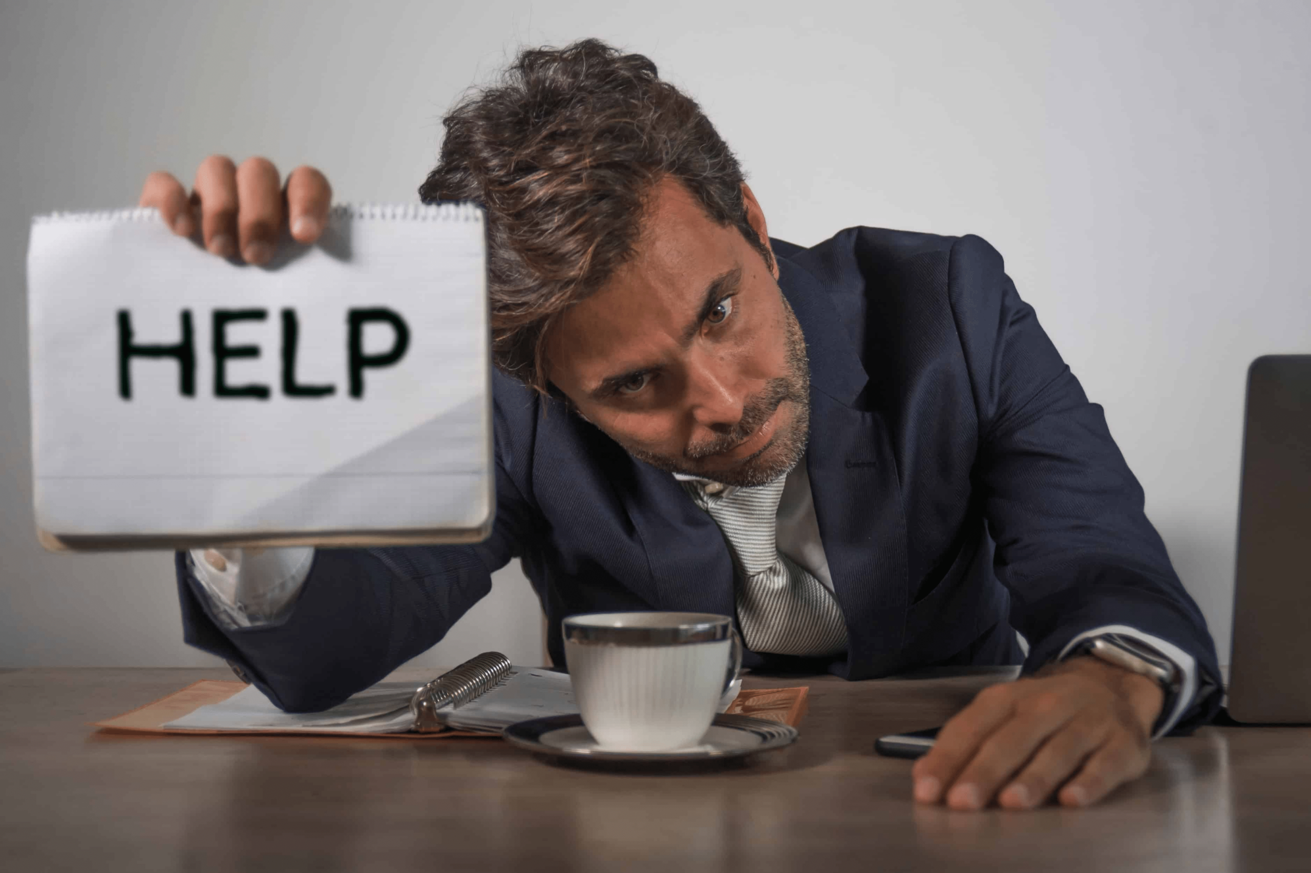 A man in an office in a suit with one hand on the desk and another holding up a help sign. His coffee computer and notebook are also on the desk as he looks depressed.