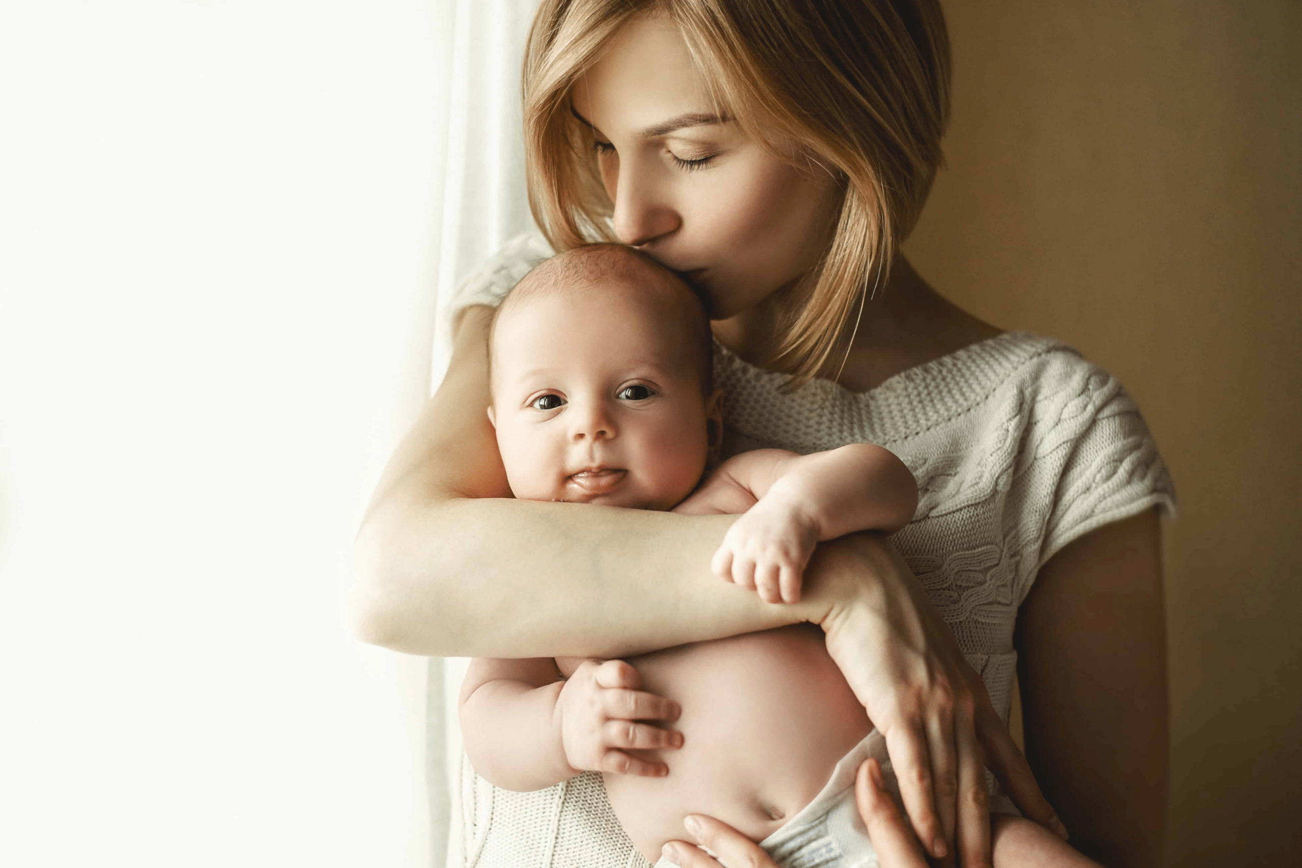 A woman holding her baby giving her baby a kiss while she stands, the baby is staring at the camera looking with both eyes open.