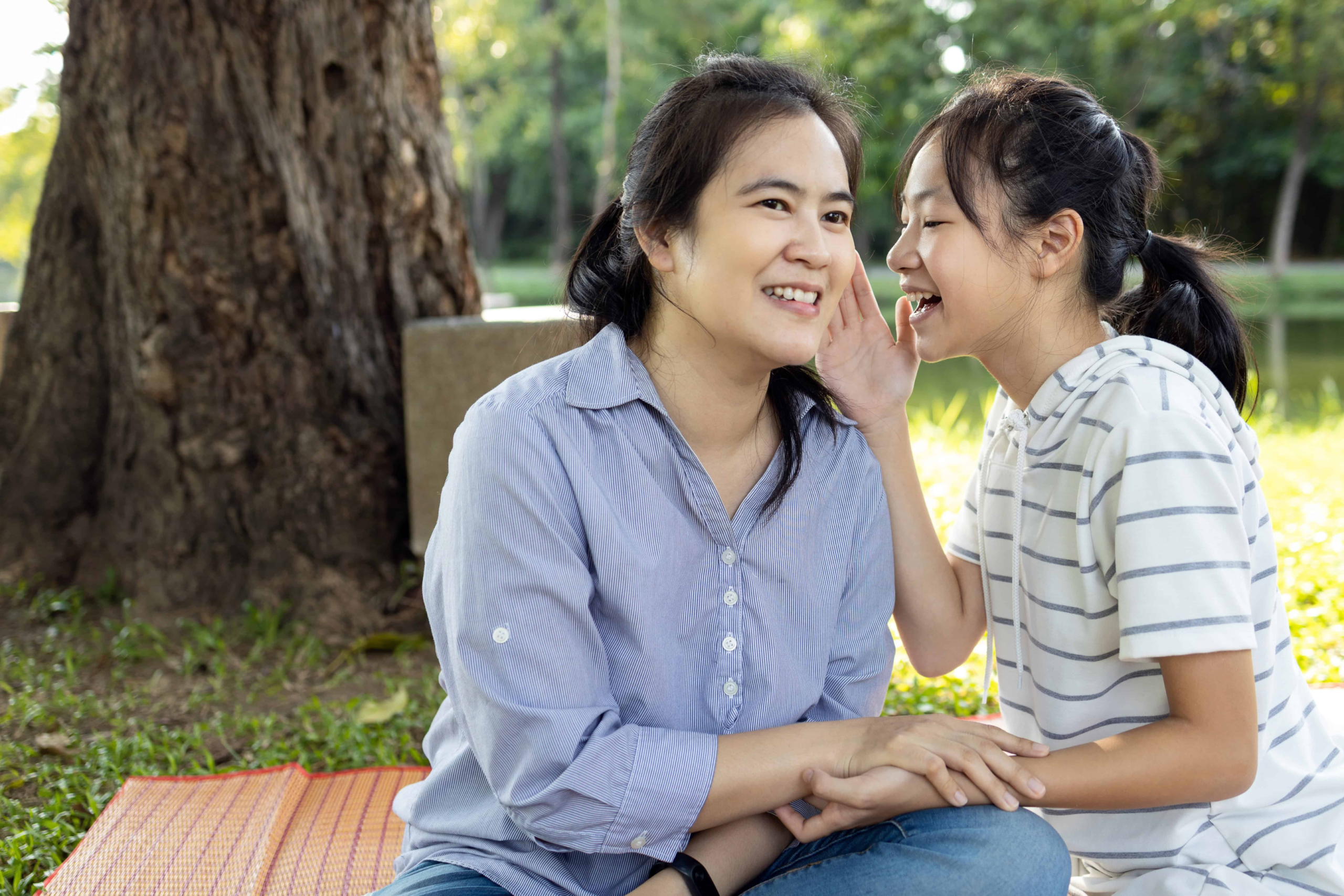 A mom and a daughter sitting in the park and listening to each other whisper secrets back and forth to each other.