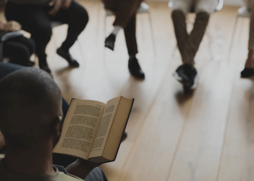 A black man sitting in front of a large crowd reading words from a book outloud.