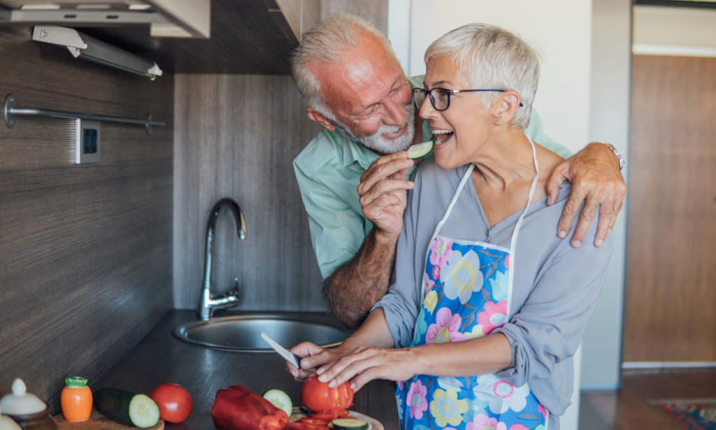 An older couple in the kitchen together, the husband is feeding his wife a cucumber.