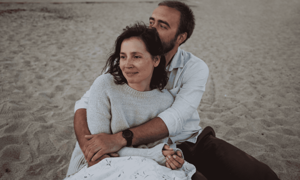 white couple holding each other tightly while sitting on the beach gazing at the sunset.
